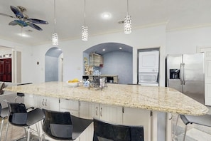 A kitchen with a large island, bar stools, a ceiling fan, pendant lights, and a refrigerator. - Sandcastle Retreat - Heart of Galveston, Dogs OK (Galveston)