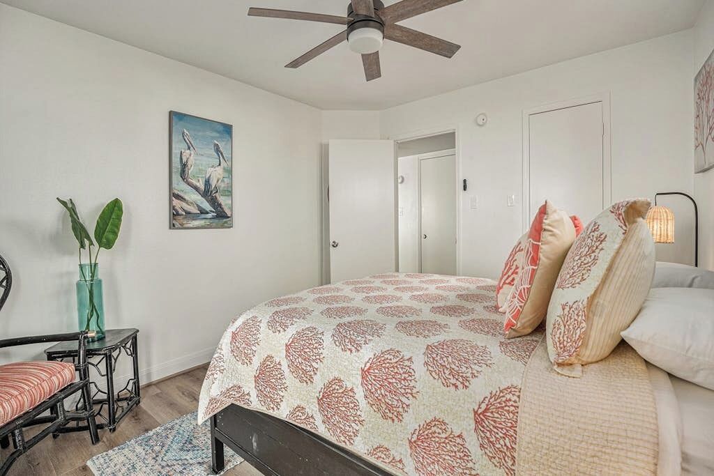 A bedroom with a bed, a ceiling fan, a door, a painting, and a small table with a plant.