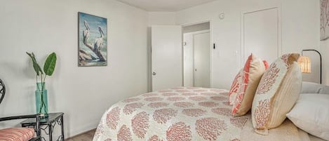 Un dormitorio con cama, ventilador de techo, puerta, un cuadro y una pequeña mesa con una planta.