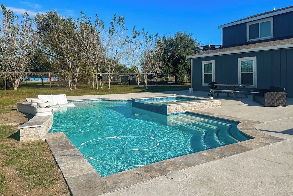 A swimming pool with a stone border and steps leading into it, surrounded by a grassy yard and trees.