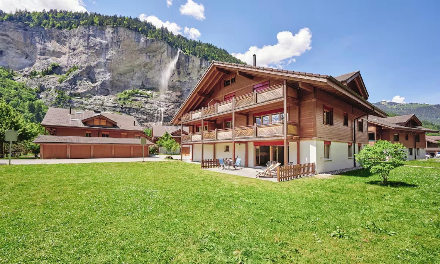 A chalet-style building with a balcony, a grassy yard, and a mountain backdrop.