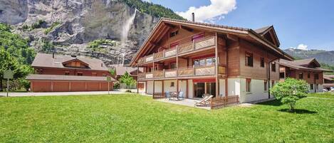 A chalet-style building with a balcony, a grassy yard, and a mountain backdrop.