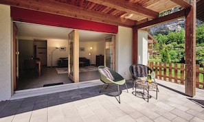 Un espacio al aire libre cubierto con una pérgola de madera, una zona de estar con dos sillones, una mesa con una botella y un vaso, y vistas a un paisaje montañoso.