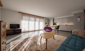 Una sala moderna con un sofá, una mesa de centro de madera y una butaca púrpura. Hay un televisor sobre un mueble de madera y una zona de cocina al fondo.