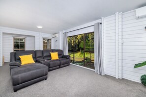 Un saló modern amb un sofà gris, una gran porta corredissa de vidre que dona a un jardí, i parets exteriors de color blanc.