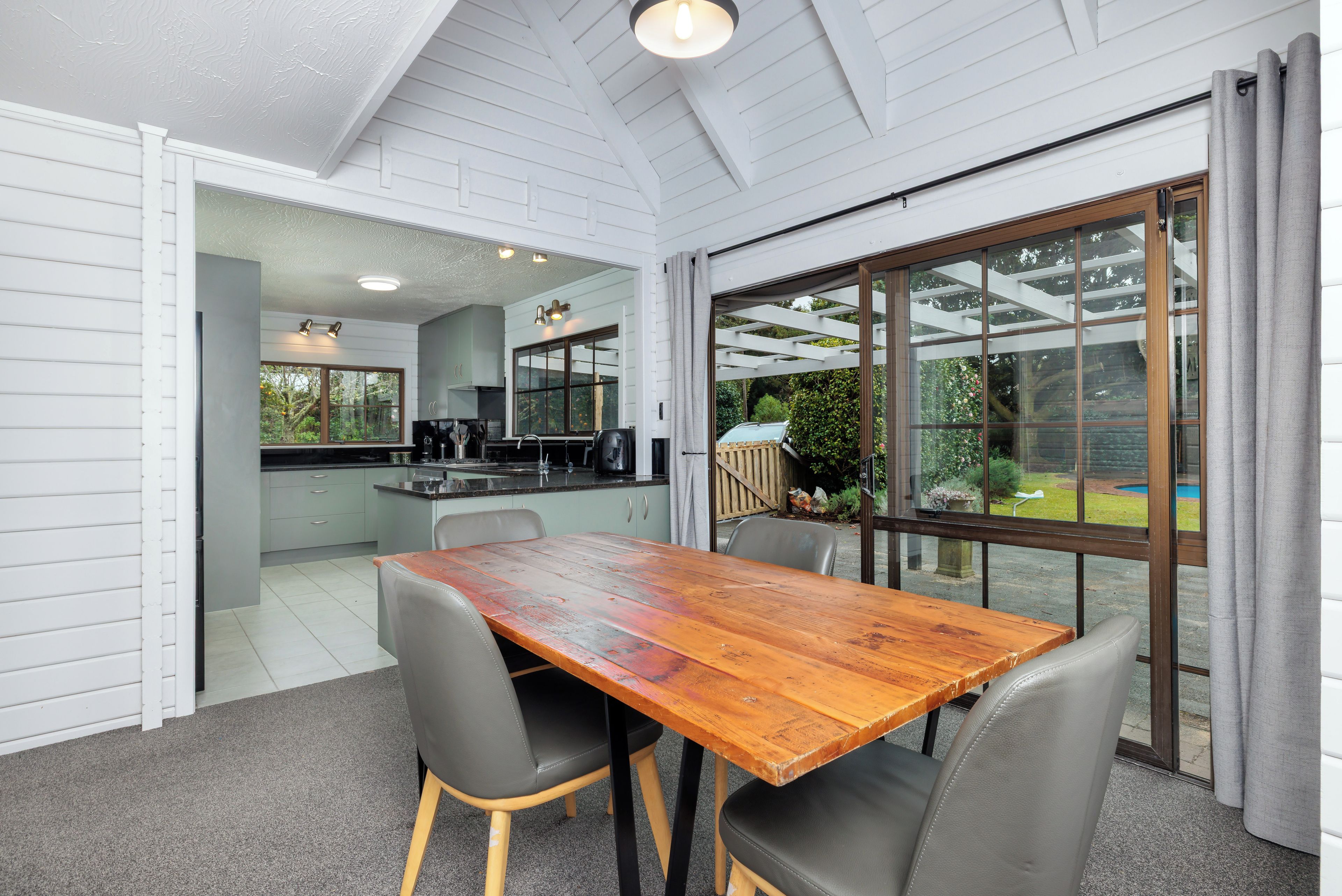A dining area with a wooden table and chairs, a kitchen in the background, and a view of a garden through large windows.