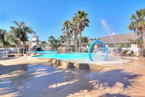 A swimming pool with a water feature, palm trees, and a building in the background. - Palm Bay 402 - Pool, Patio & Dog-Friendly (Corpus Christi)