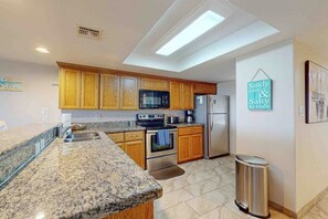 A kitchen with granite countertops, wooden cabinets, stainless steel appliances, and a trash can. - Suntide II Condominiums #101 (South Padre Island)