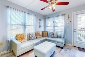 Ein modernes Wohnzimmer mit einem Ecksofa, einem Deckenventilator und großen Fenstern mit Jalousien.