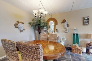 A dining area with a round wooden table, wicker chairs, a chandelier, and decorative wall art including a turtle, seahorse, and a mirror. - Beachview Condominiums #111 (South Padre Island)