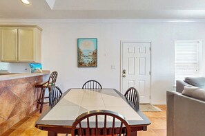 A dining area with a rectangular table, four chairs, a kitchen with cabinets, a door, and a window with blinds. - Las Puertas Condominiums 201 (South Padre Island)