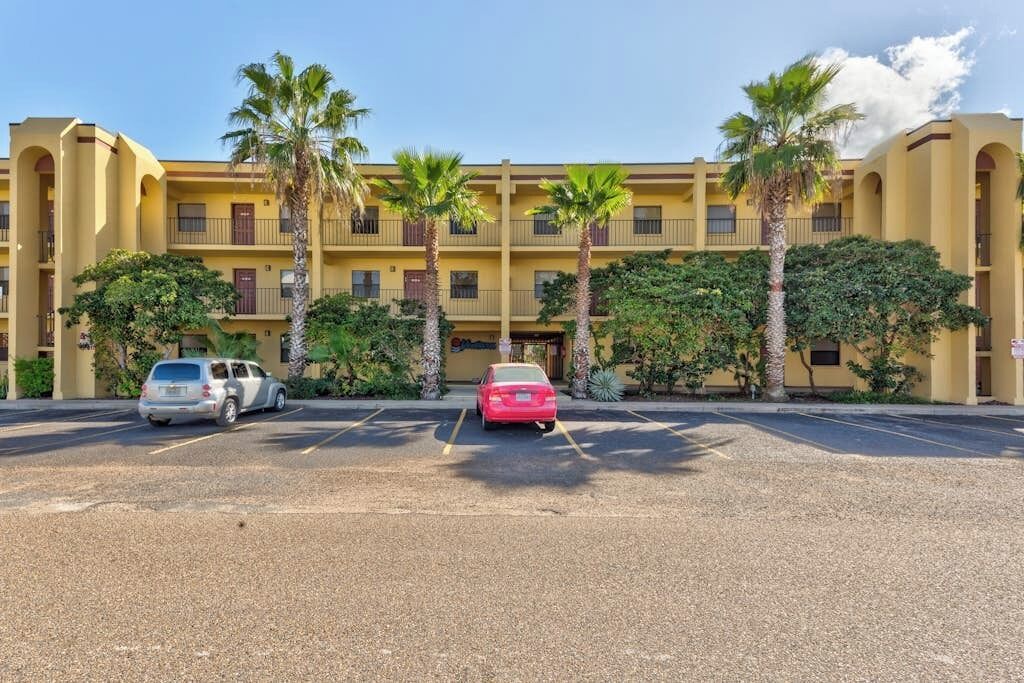 Un hôtel à plusieurs étages, avec une façade jaune, des palmiers et un parking où sont garées plusieurs voitures.