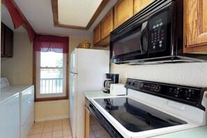 A compact kitchen with a microwave, stove, and a window with a red curtain. - Mezquite Duplex Beach House (South Padre Island)