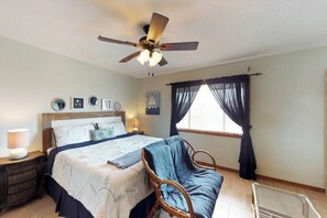 Une chambre à coucher comprenant un lit, une chaise, une table de chevet, un ventilateur de plafond et une fenêtre avec des rideaux.