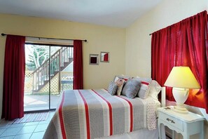 A bedroom with a bed, a nightstand, a lamp, a window with curtains, and a view of a staircase leading outside. - Mezquite Duplex Beach House (South Padre Island)