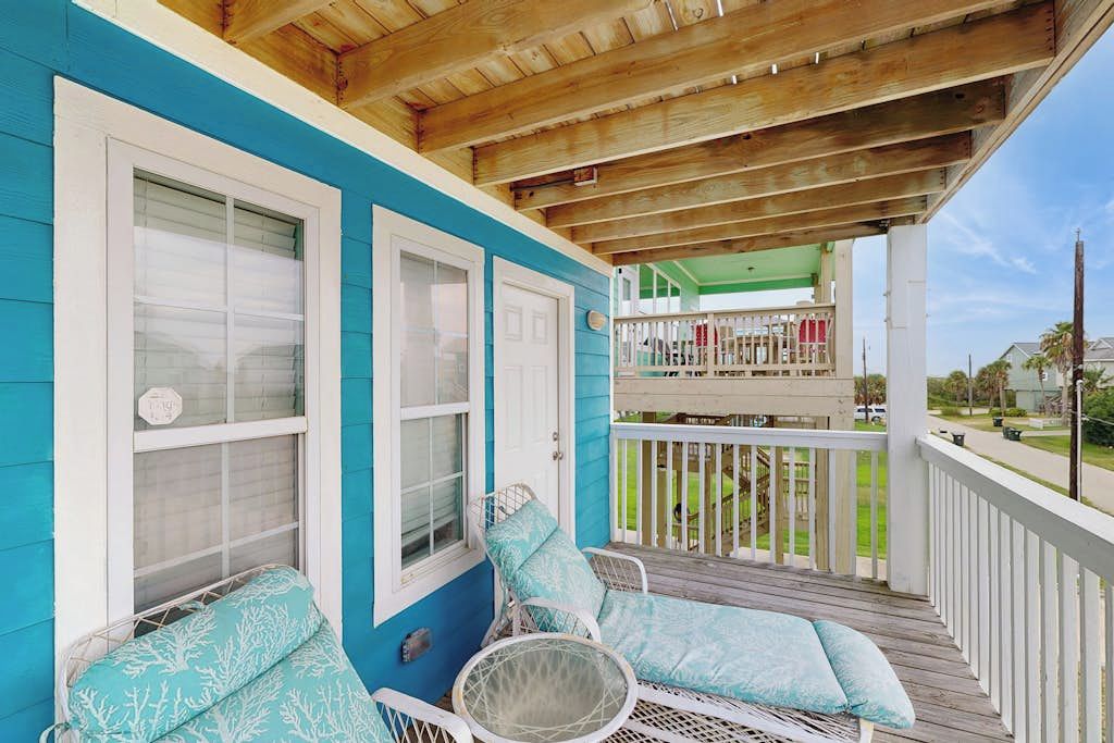 A covered porch with two white wicker chairs and a cushioned bench, a wooden railing, and a view of a green lawn and a house in the background.