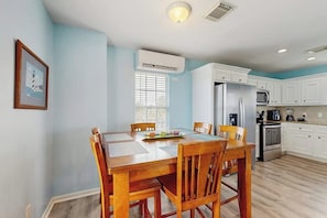 A dining area with a wooden table and chairs, a window with blinds, and a kitchen with white cabinets and appliances. - Beachwood - Fire Pit, Steps to Sand, Dogs OK (Galveston)