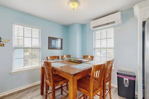A dining area with a wooden table and chairs, two windows with blinds, an air conditioning unit, and a trash bin. - Beachwood - Fire Pit, Steps to Sand, Dogs OK (Galveston)