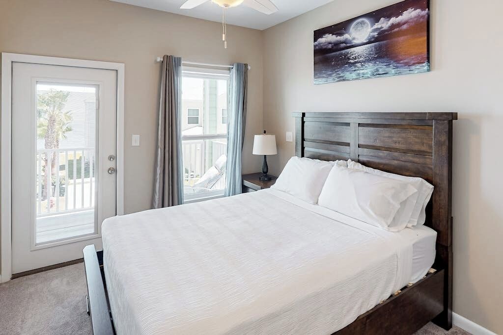 A hotel room with a bed, a nightstand, a lamp, a painting, and a view of the outside.