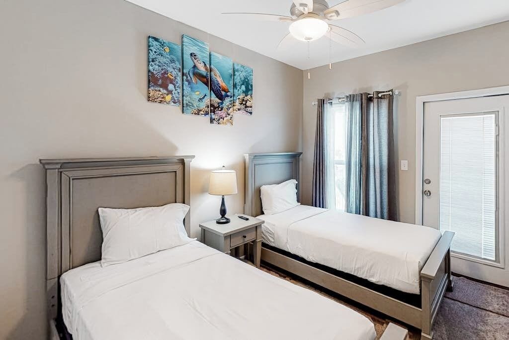 Une chambre d’hôtel avec deux lits, un ventilateur de plafond, des œuvres d’art accrochées au mur et une porte avec des rideaux.