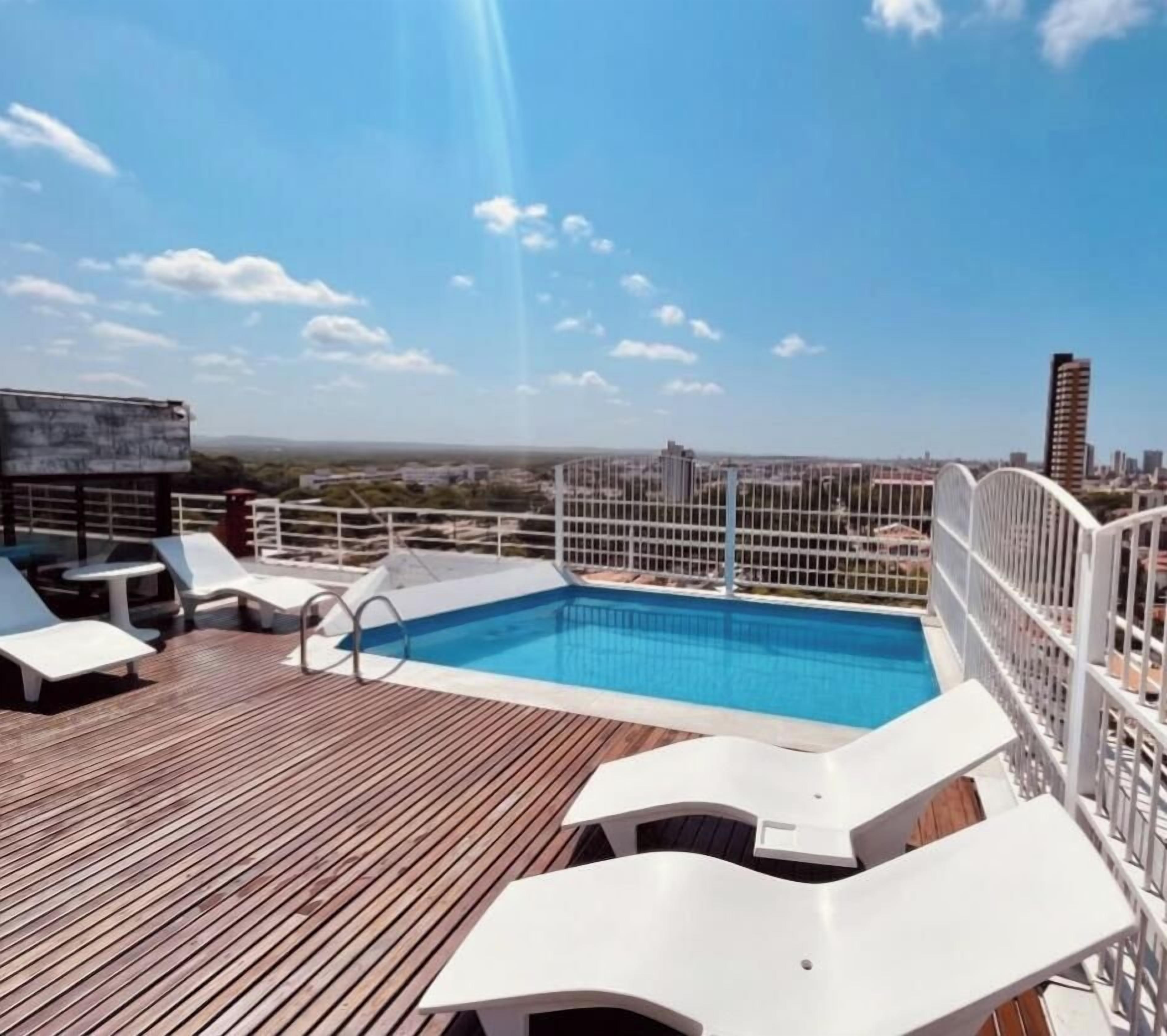 Área de piscina no terraço com vista para o horizonte urbano, cercada por grades brancas e espreguiçadeiras.