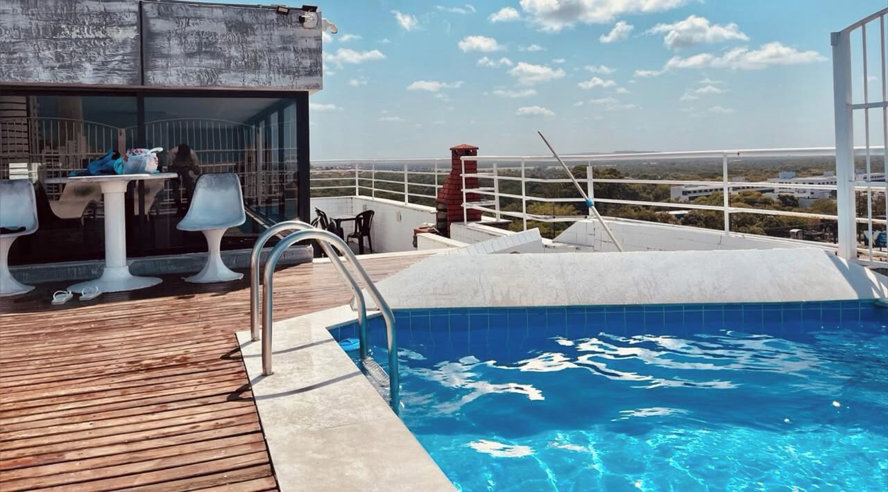 A rooftop swimming pool with a wooden deck, a table with chairs, and a view of the landscape.
