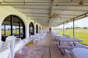 Une terrasse extérieure couverte, dotée de chaises et de tables en plastique blanc, de grandes fenêtres et d’un ventilateur de plafond.