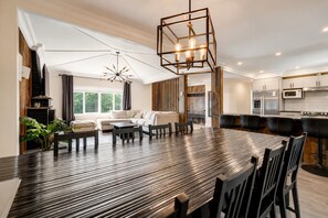 Una cocina moderna con electrodomésticos de acero inoxidable, un comedor con una larga mesa de madera y una sala de estar con un sofá y un ventanal.