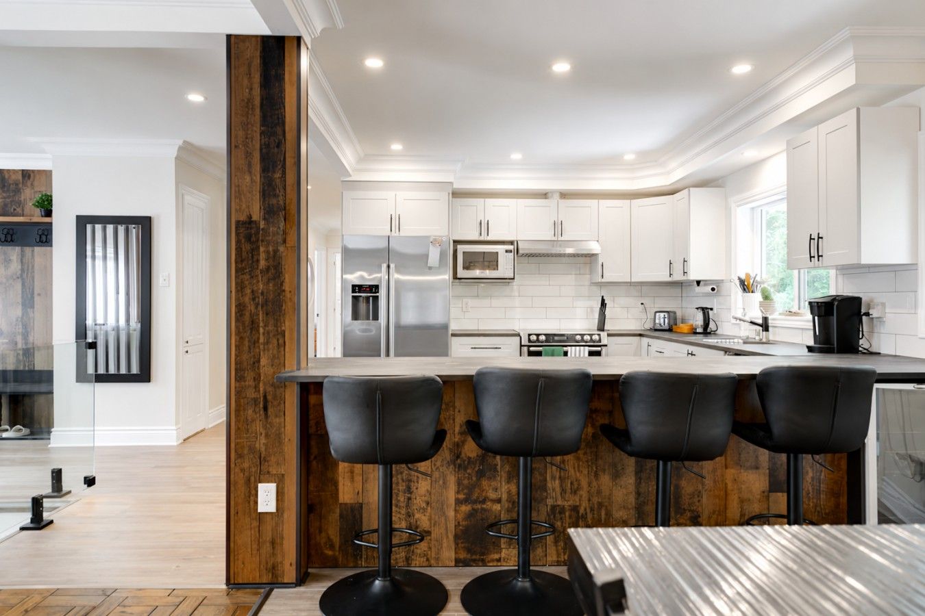 A modern kitchen with white cabinetry, a central island with bar stools, stainless steel appliances, and a wooden accent wall.