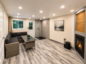 A modern living room with a sofa, a glass-top coffee table, a fireplace, and a large window.