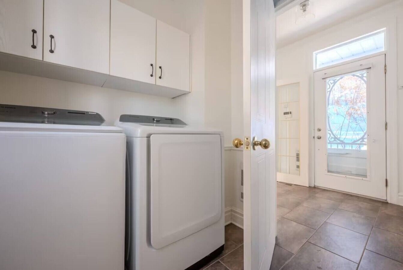 A laundry room with two white appliances, a door leading to a balcony, and a view of a tree through a glass door.