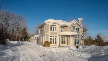 A large, two-story house with a balcony, surrounded by snow-covered ground and trees.