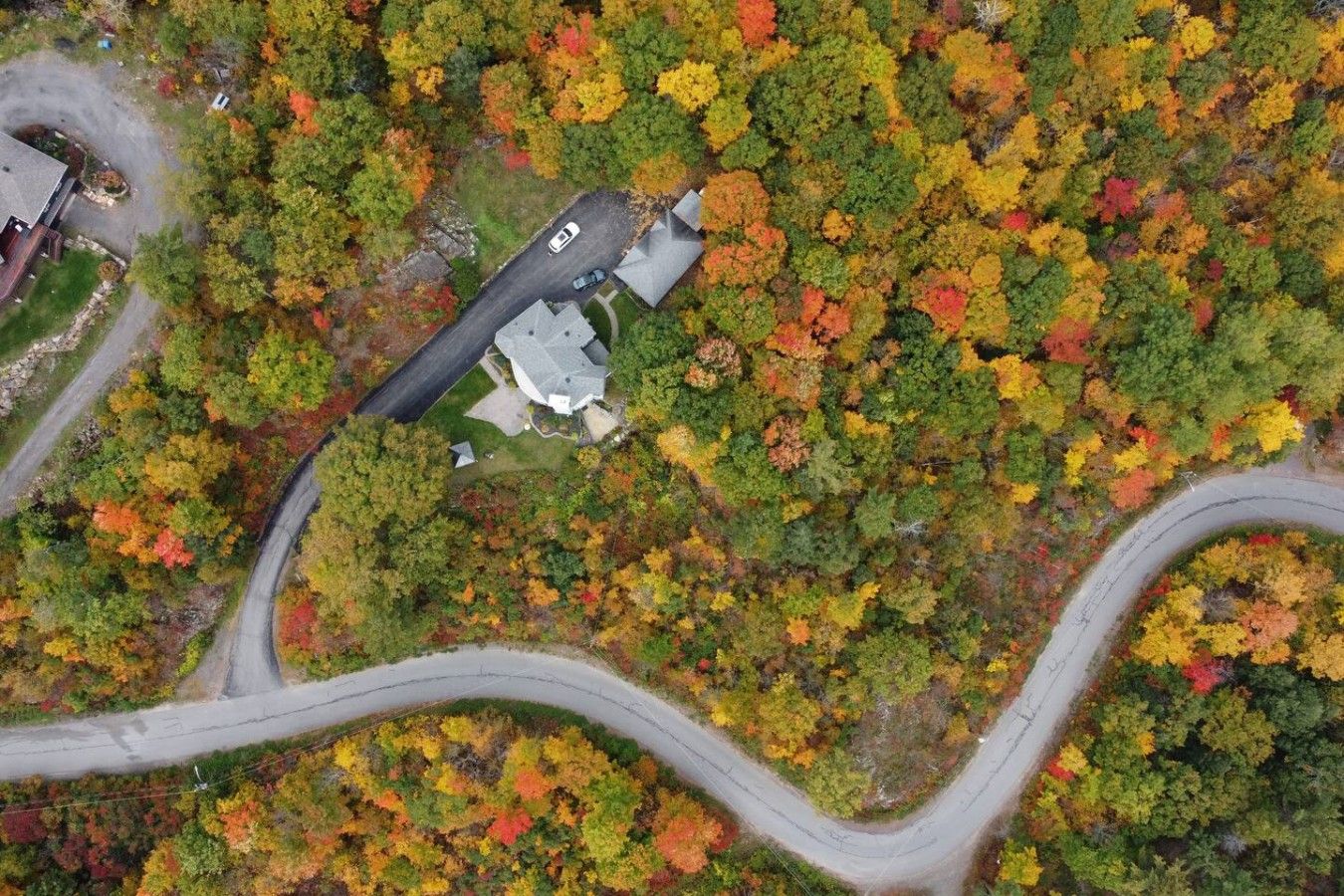 An aerial view of a residential area with a winding road, a house with a sloped roof, and dense surrounding trees.