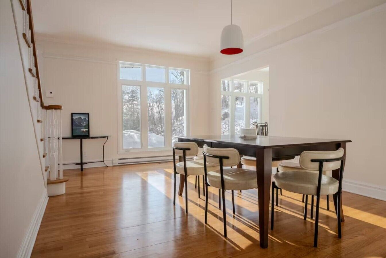 A dining area with a wooden table and chairs, large windows, and a staircase.