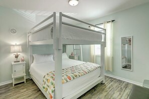 Una cama litera con estructura blanca y cubrecama estampado con piñas. Hay una mesita de noche con una lámpara y un objeto decorativo pequeño. Un espejo está montado en la pared a la derecha de la cama.
