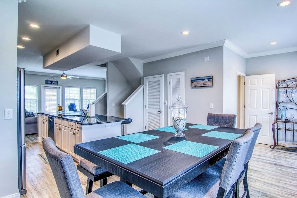 Un coin repas avec une table dressée pour quatre personnes, une cuisine en arrière-plan et un escalier menant à un autre niveau.