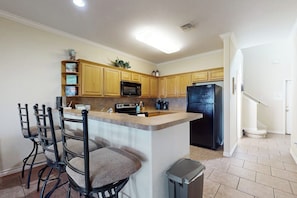 A kitchen with wooden cabinets, a black refrigerator, a microwave, a sink, and a trash can. There are three bar stools with cushioned seats. - Village Townhomes on the Greens Unit 106 (Corpus Christi)