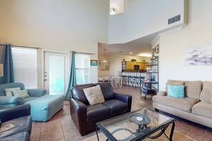 A spacious living room with a sofa, armchair, and coffee table, a kitchen in the background, and a dining area with chairs. - Village Townhomes on the Greens Unit 106 (Corpus Christi)
