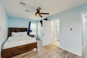 A bedroom with a wooden bed, a ceiling fan, and a door leading to a bathroom. - Blessings on the Bay (Corpus Christi)