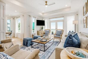 A spacious living room with a large window, a flat-screen TV, and a ceiling fan. - Beach Haus - Ocean Views, Sleeps 22 & Bunk Room (Galveston)