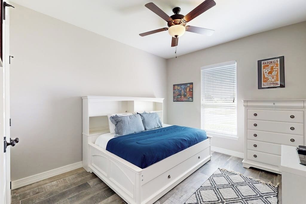 Une chambre à coucher avec un lit blanc, un ventilateur de plafond, une fenêtre avec des stores, une commode et un tapis.