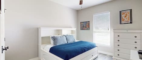 A bedroom with a white bed, a ceiling fan, a window with blinds, a dresser, and a rug.