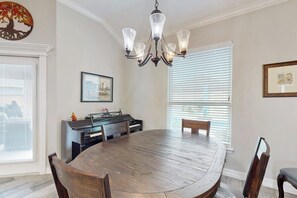 Un espace repas comprenant une table ronde en bois, des chaises, un lustre, un piano et des œuvres d’art encadrées accrochées aux murs.