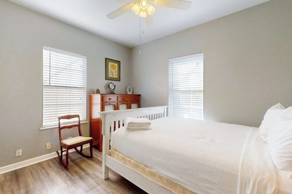 A bedroom with a bed, a chair, a dresser, and two windows with blinds.