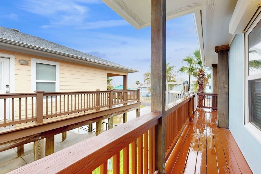 Een houten terras met een balustrade, een gebouw met een lichtgekleurde gevel en een heldere lucht.