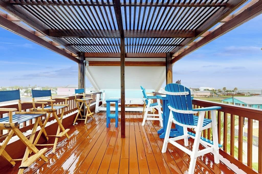A covered outdoor deck with wooden flooring, blue and white Adirondack chairs, and a table.