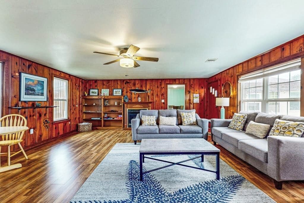 A living room with wooden walls, a ceiling fan, a grey sofa, a coffee table, and a fireplace.