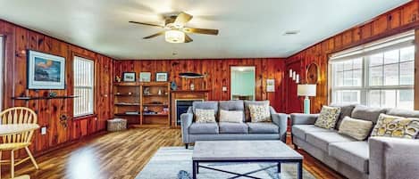 A living room with wooden walls, a ceiling fan, a grey sofa, a coffee table, and a fireplace.