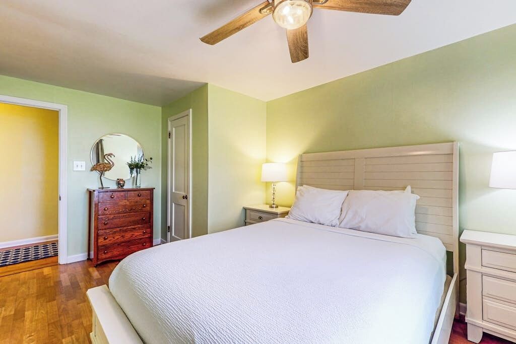 A bedroom with a bed, a wooden dresser, a nightstand with a lamp, and a ceiling fan.