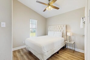 Ein Schlafzimmer mit einem Bett, einem Deckenventilator, einem Nachttisch mit Lampe und einem Fenster mit Jalousien.